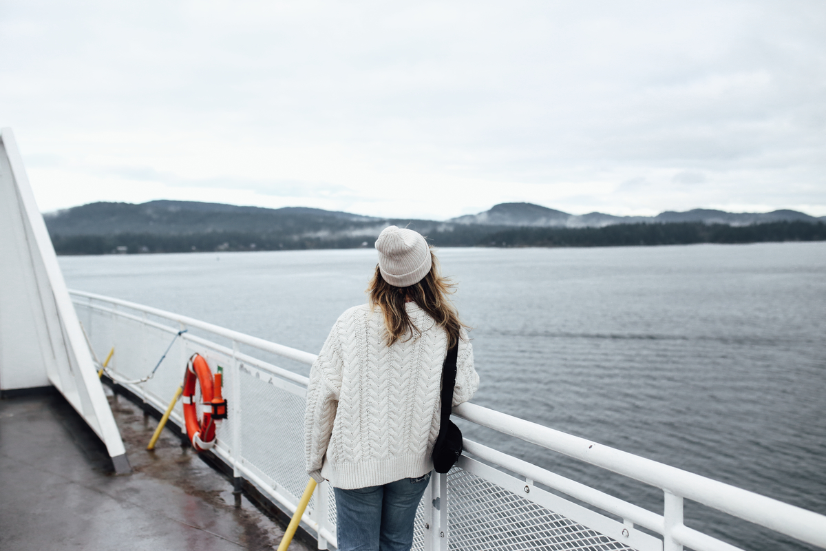 A woman looks out from the ferry deck at the ocean. She is wearing a white sweater and matching toque. The weather is overcast.