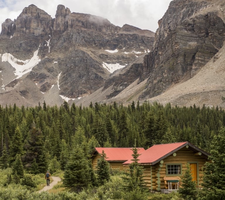 A hiker walks past by Assiniboine Lodge cabins - a log cabin with red roof surrounded by a mountain backdrop.