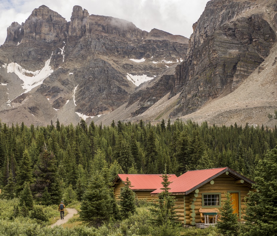 A hiker walks past by Assiniboine Lodge cabins - a log cabin with red roof surrounded by a mountain backdrop.