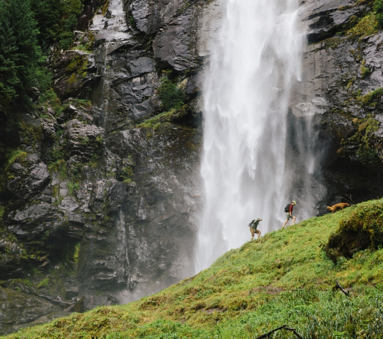 Two hikers walk beside Exstew Falls, it drops over a rocky ledge. They are hiking up a steep green ridge.