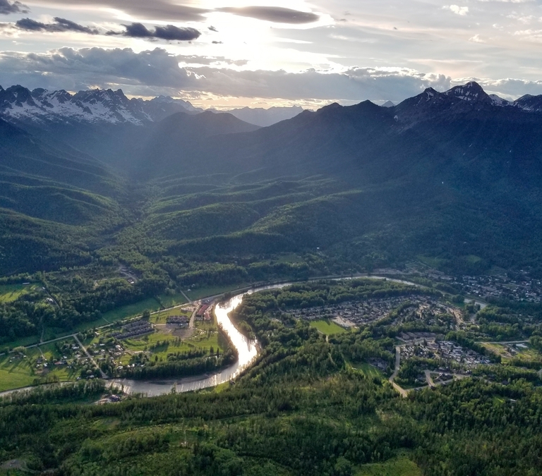A scenic view of the mountains and valley below. A river runs through a lush green fields. The sun is starting to set behind the mountains.