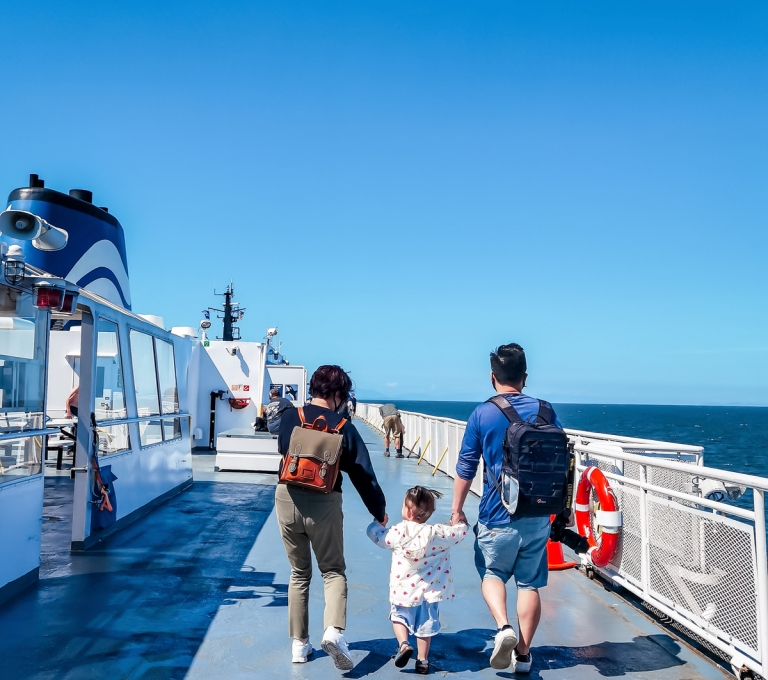 A family walks along the top deck of a BC Ferries boat on a sunny blue ski day. Two parents hold the hand of small child between them.