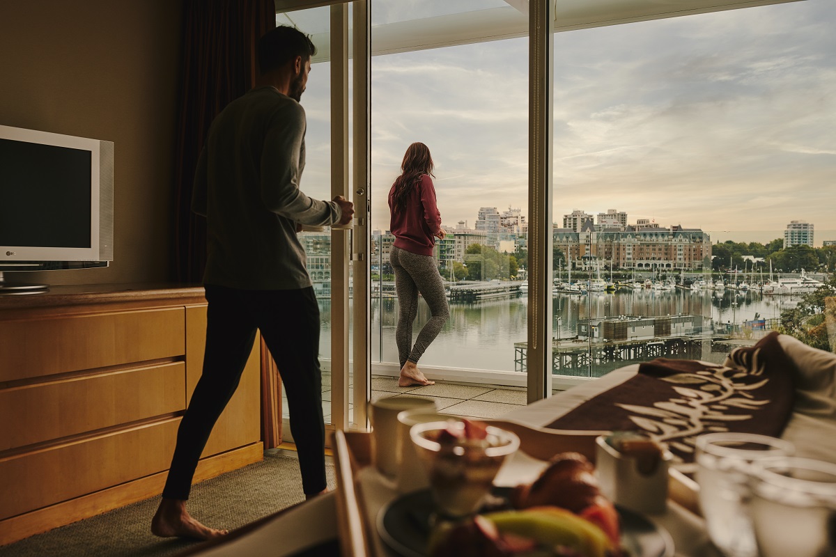A couple in a room at the Inn at Laurel Point. One person stands outside on the balcony looking out at the harbour, the other person walks towards the patio doors.