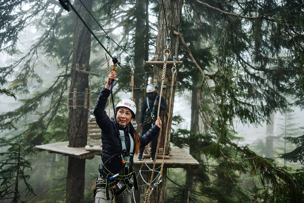 Woman on a ropes course at Grouse Mountain - a popular Vancouver attraction. She is wearing a white helmet and holding a line above her head and walking carefully across the ropes below.