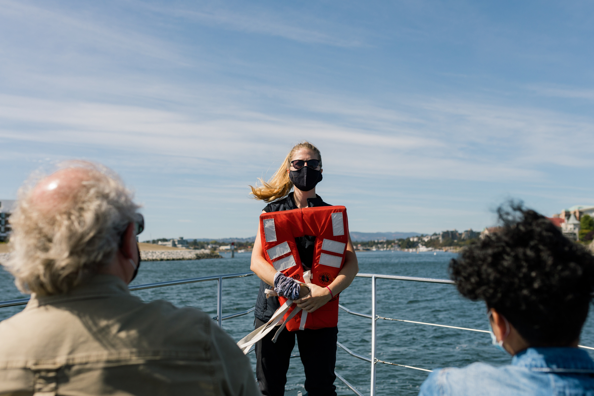 An employee of Springtide Whale Watching holds a red lifejacket facing the crowd on the boat deck. She is doing a safety briefing to passengers.