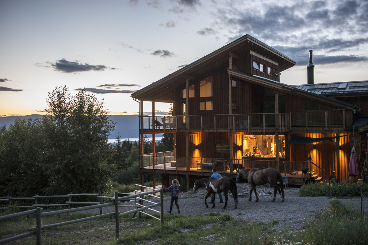 Two people walk horses back to their pastures outside the Myra Canyon Lodge in Kelowna. The sun is setting, and the lights are on inside the lodge.