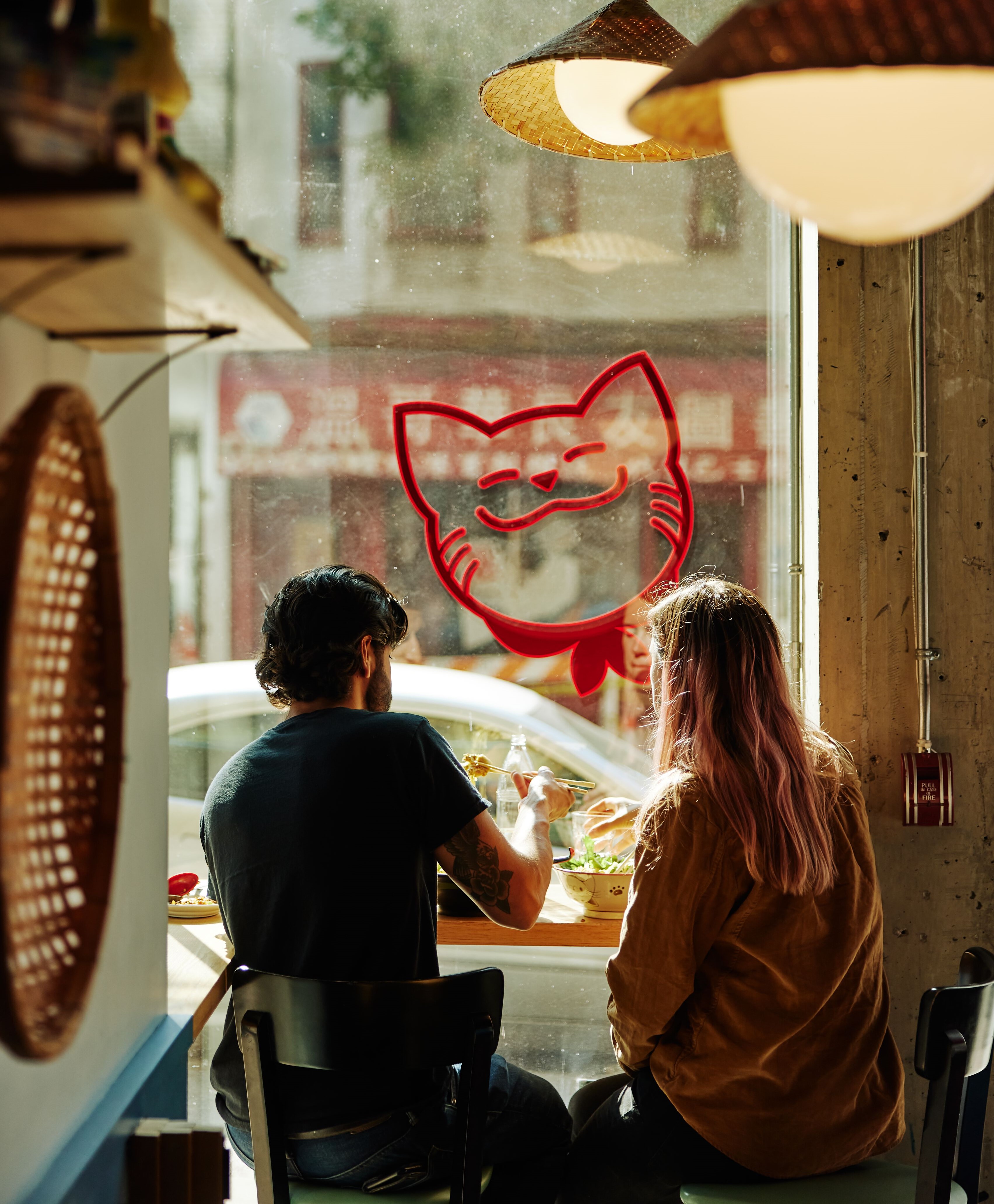 Two diners sit on stools at a window at Fat Mao Noodles in Vancouver's Chinatown. There is a decal of a cat outline in red on the window in front of them.