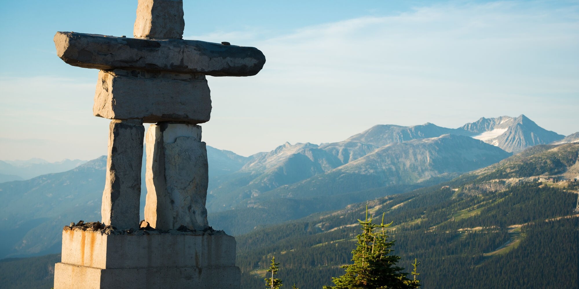 In the foreground on the left side of the frame is a giant Inukshuk statue. In the distance are forested mountains.