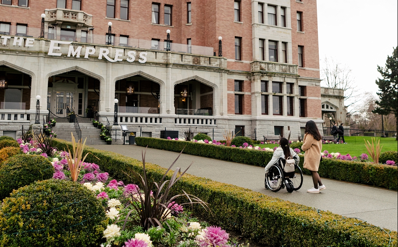 Two people approach the Fairmont Empress in Victoria. Flower gardens are seen in the foreground.