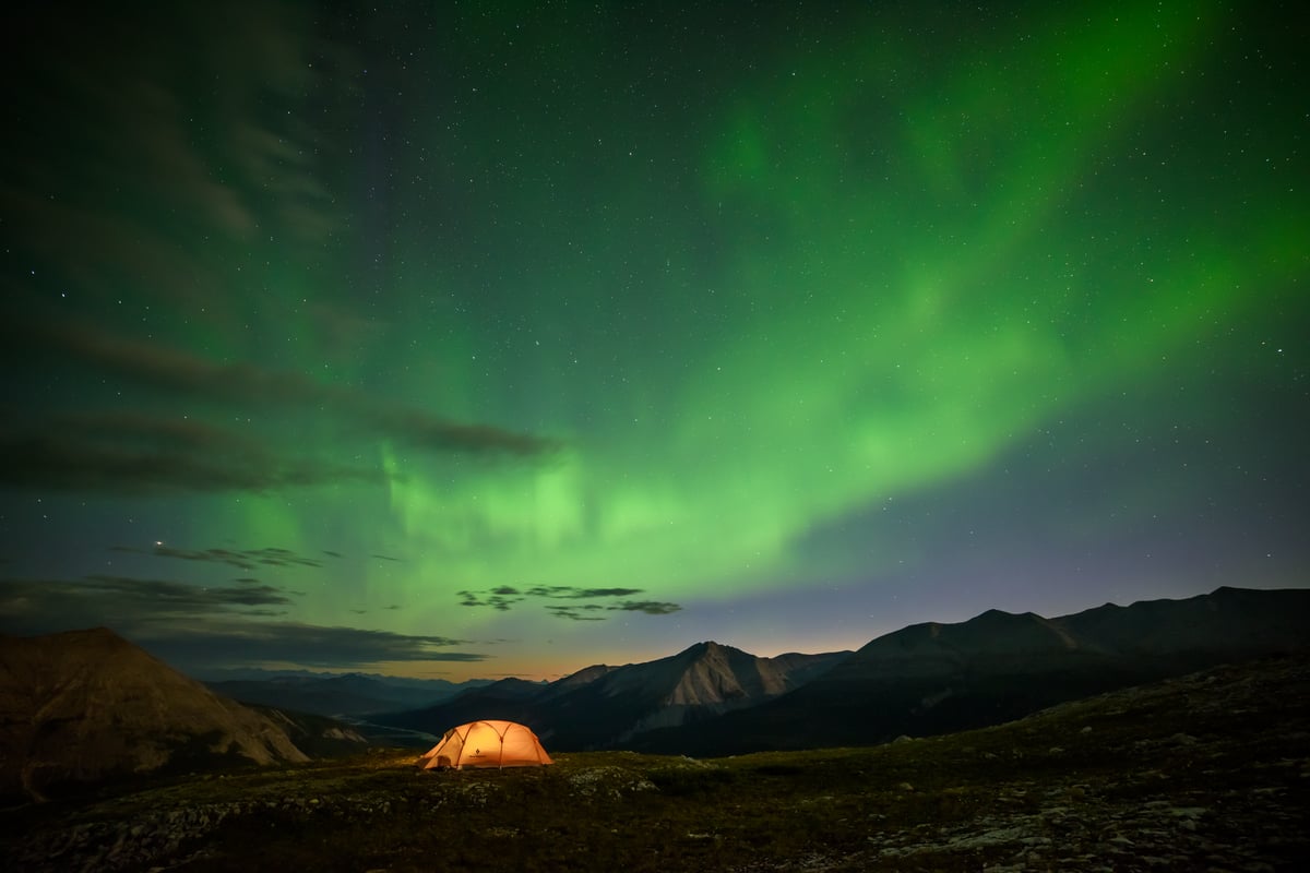 The Northern Lights are seen over the mountains. A single orange tent is lit up.