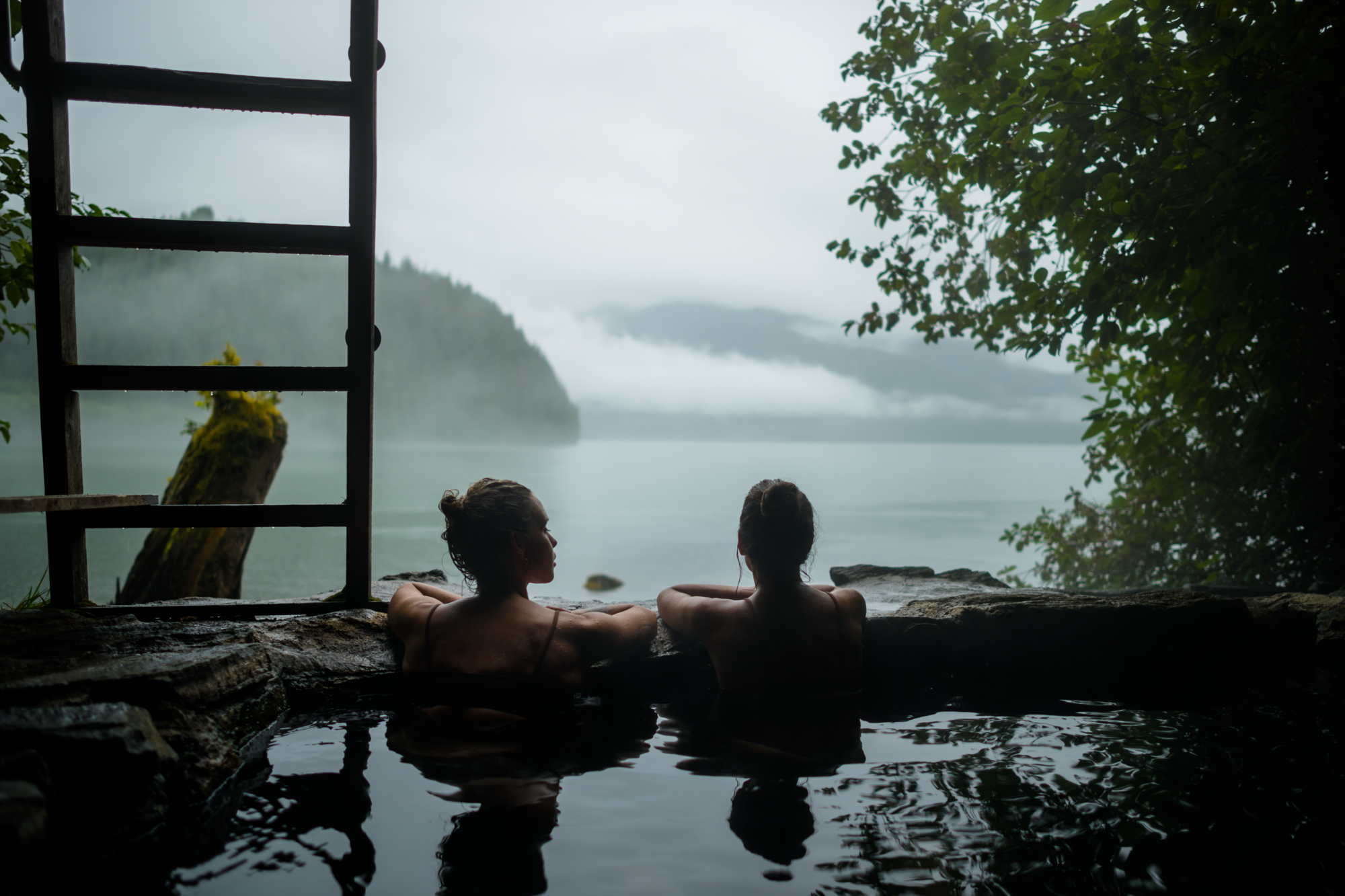 Two women look out from a hot spring over the ocean. It is dark and overcast.