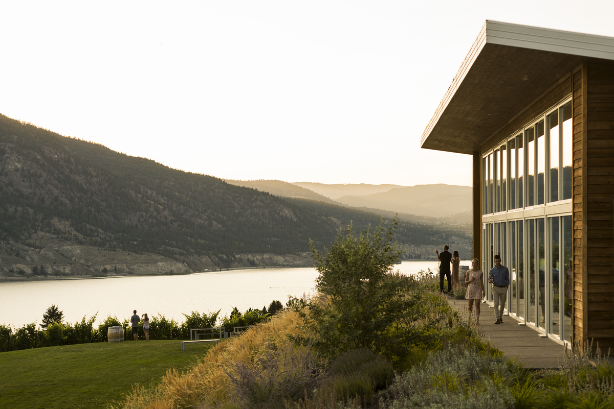 Outside shot of the Poplar Grove Winery with people walking alongside the glass walls. The winery overlooks a lake.
