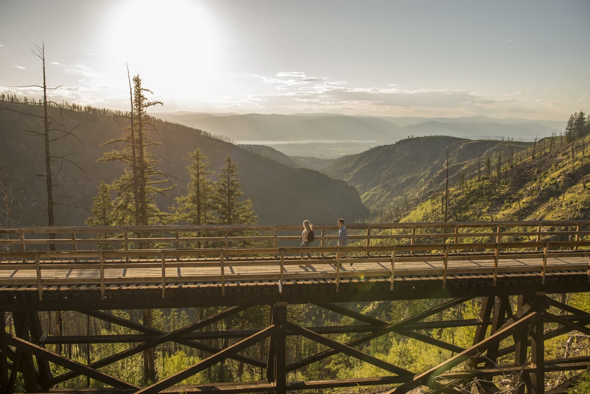A person looks out from a trestle bridge in the Myra Canyon towards the mountain range. The sun is high in the sky and shines down on the green mountains below.