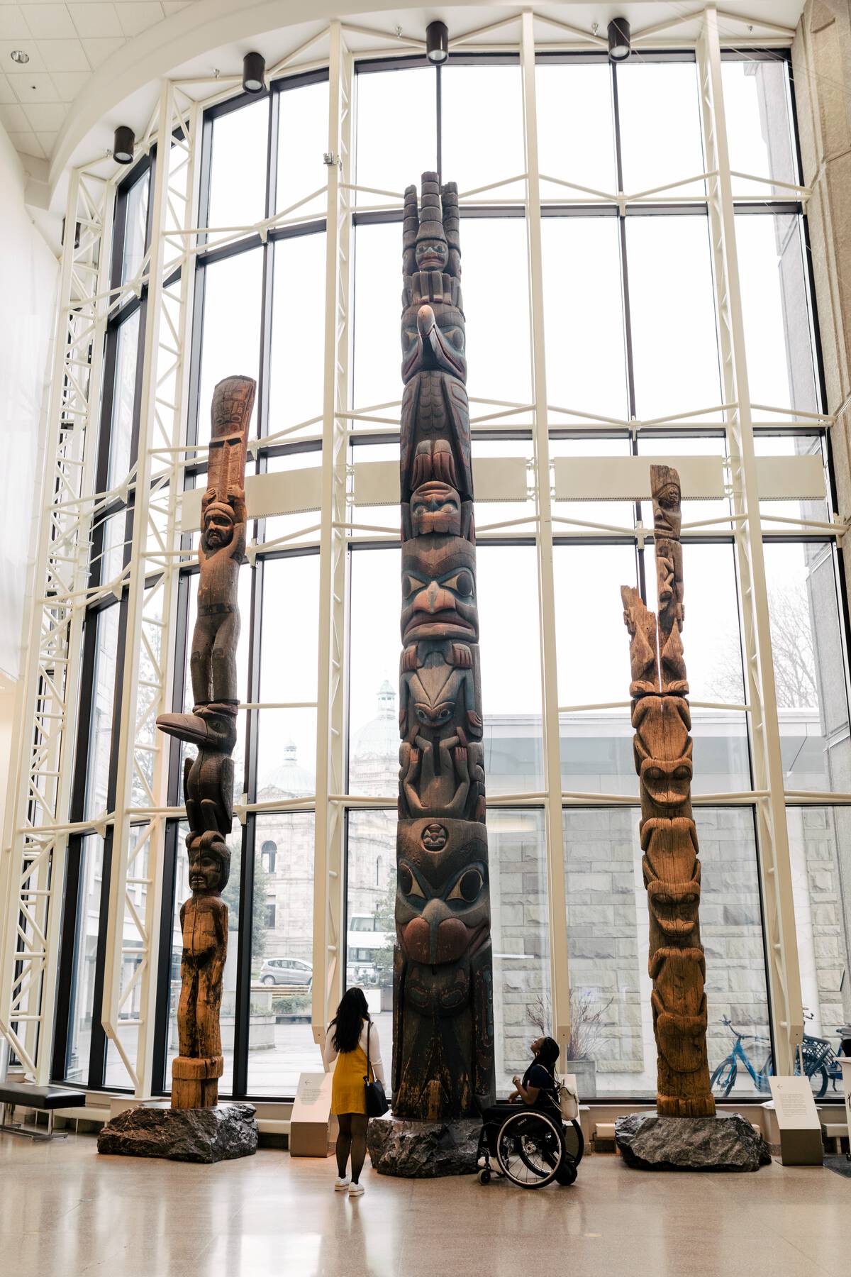 Two people look up at a collection of three totem poles at the Royal BC Museum. 