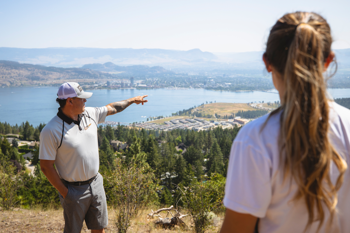 A man points out in the distance towards the lake on a walking tour of the Moccasin Trails. A woman with a pony tail is in the foreground listening.