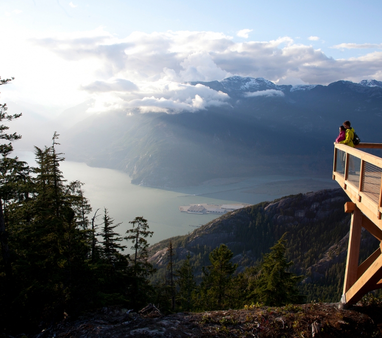 View of Howe Sound from the top of the Sea to Sky Gondola. Ocean and mountains are in the distance, and evergreen trees are in the foreground.