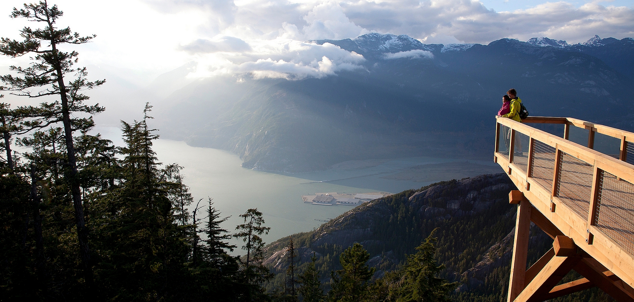 View of Howe Sound from the top of the Sea to Sky Gondola. Ocean and mountains are in the distance, and evergreen trees are in the foreground.