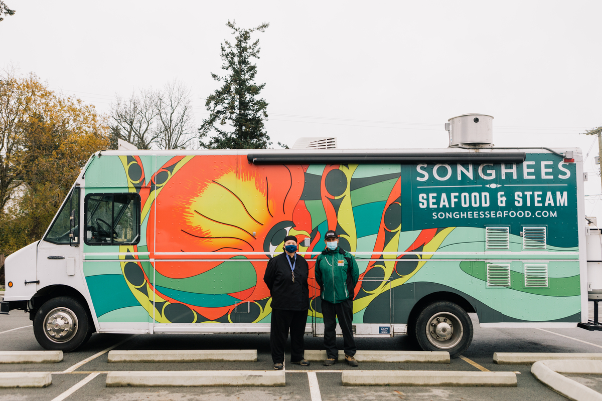 Two people stand in front of the Songhees Seafood & Steam food truck. The truck is painted with bright orange and green colours.