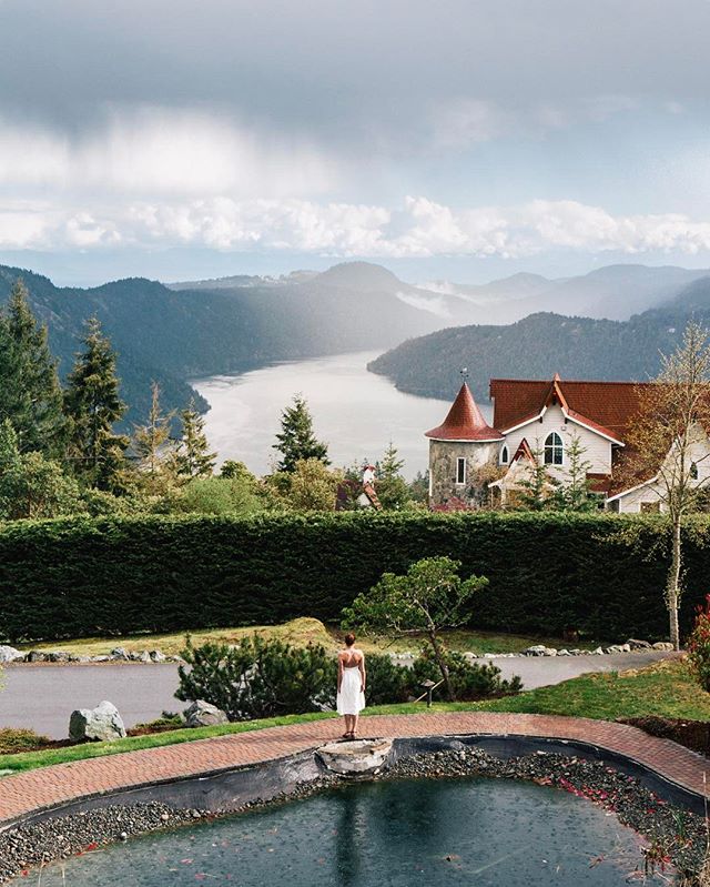 A woman stands on a red brick path taking in the views over the lake and mountains from Villa Eyrie Resort. There is a small pond behind here. The resort has a red roof and white walls.