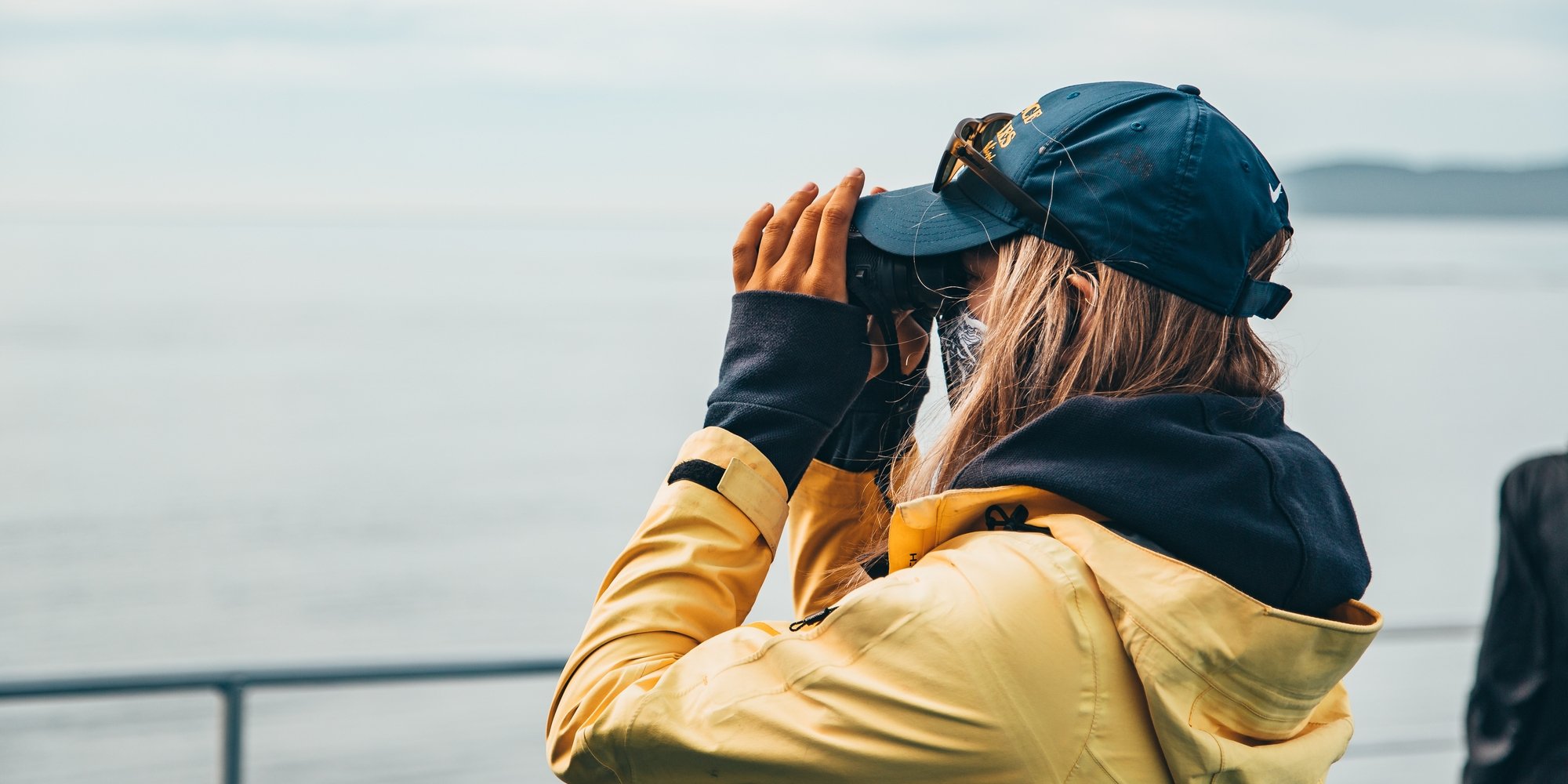 A guide wearing a yellow rain jacket and a blue cap on a Prince of Whales tour scans the water through a pair of binoculars