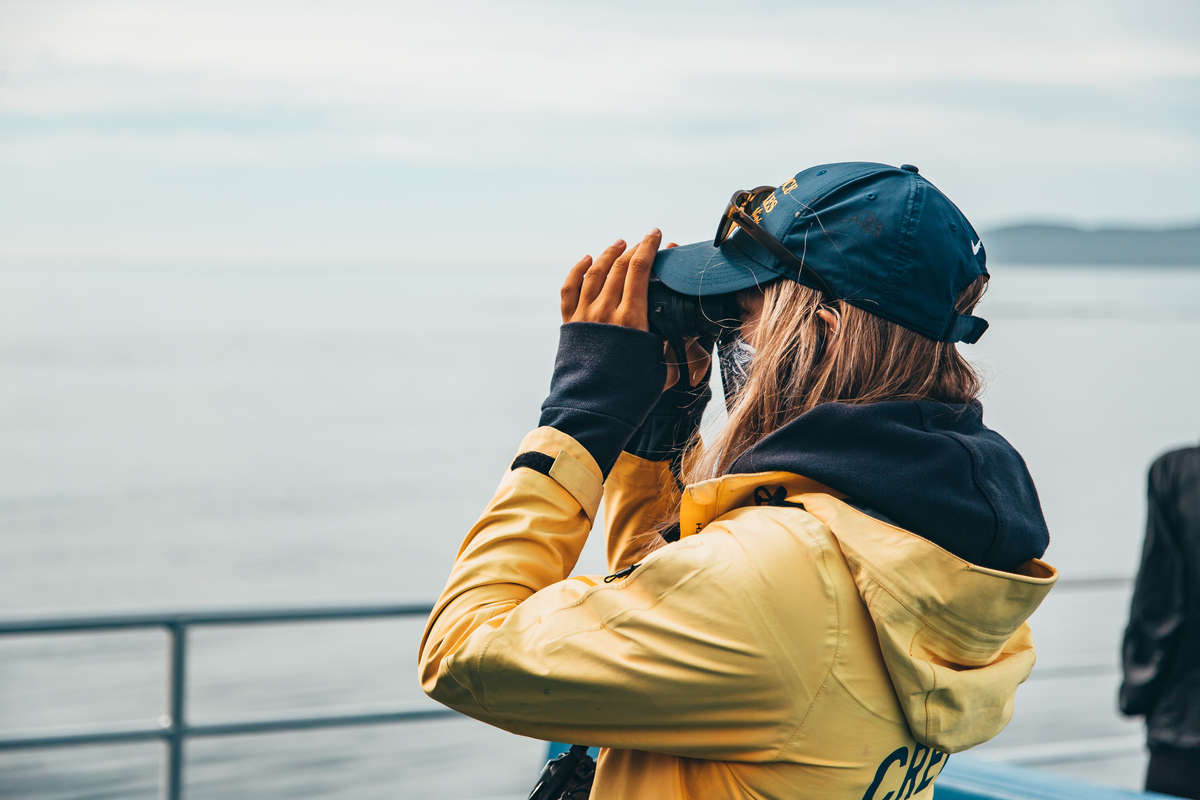 A whale watching guide in a yellow jacket and blue baseball cap looks out through binoculars to the ocean.