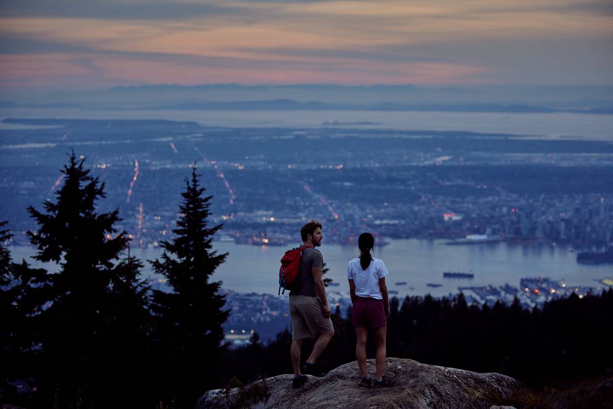 From an outlook over the city, two people look down at Vancouver. It's the evening time and the lights are just turning on across the city.