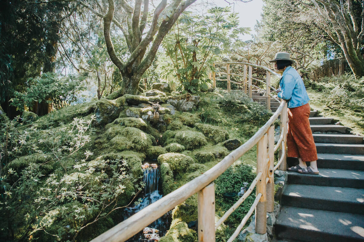 Woman in a blue shirt, rust-coloured pants and a hat stands against a railing on a staircase overlooking trees, shrubs and flowers