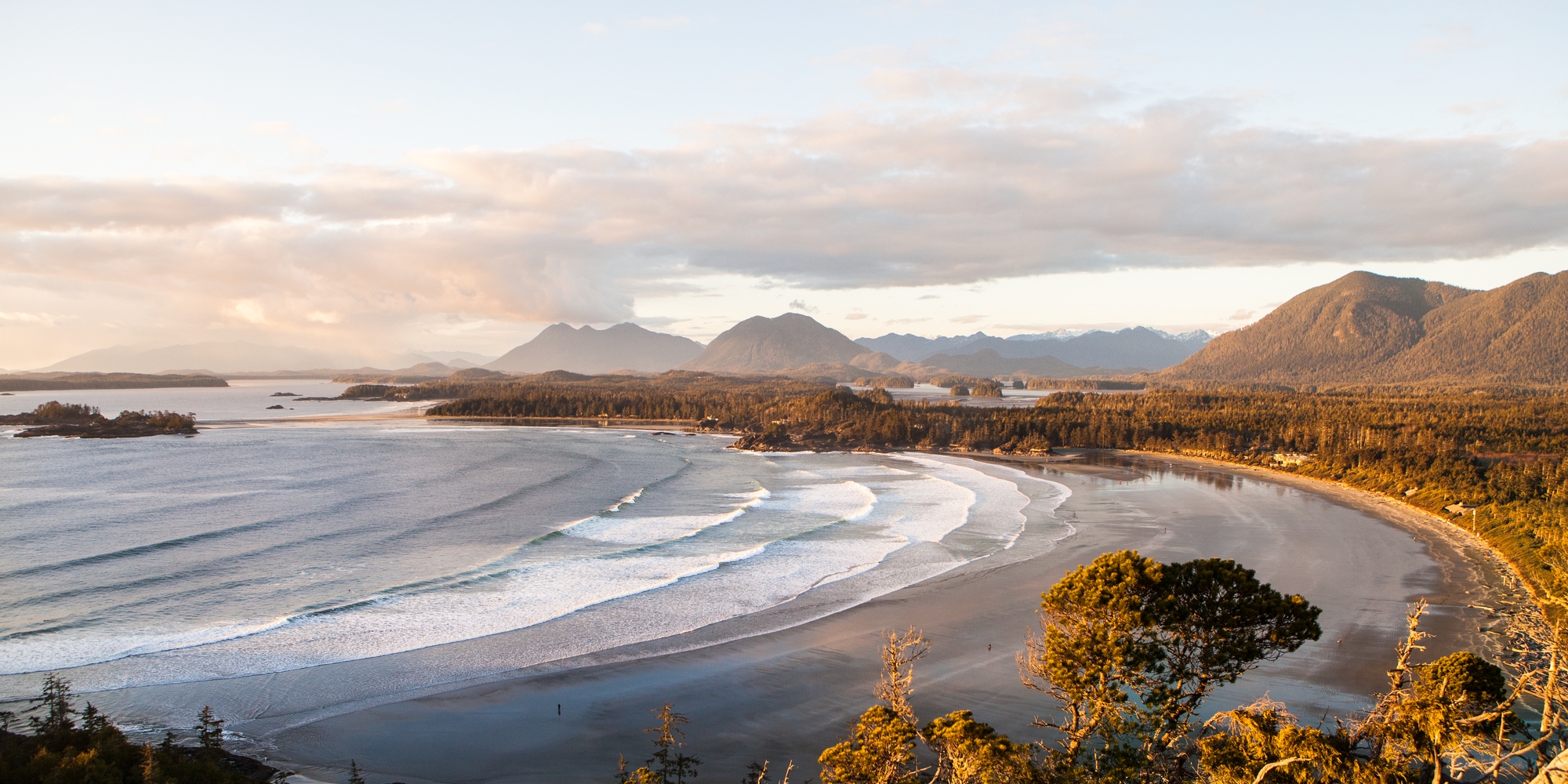 The waves roll onto shore with mountains in the background of View of Cox Bay in Tofino