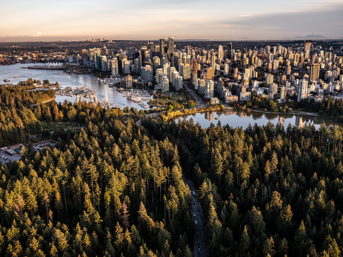 Aerial shot of downtown Vancouver with Stanley Park in the foreground. The sun is setting lighting up the sky in light pink and blues. A large cluster of tall office buildings are in the downtown area.