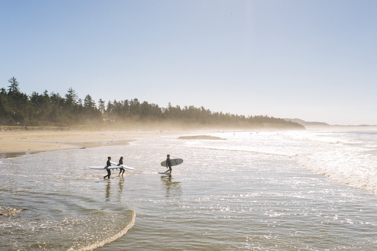 Three people wade into the ocean with surf boards on a sunny day in Tofino.