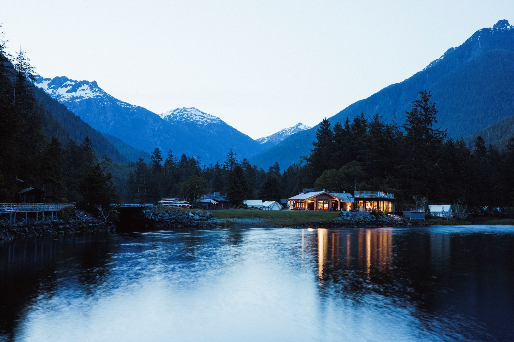 A nighttime view of the Clayoquot Wilderness Lodge. The lights of the lodge are reflected on the water and snowcapped mountains are seen in the distance.
