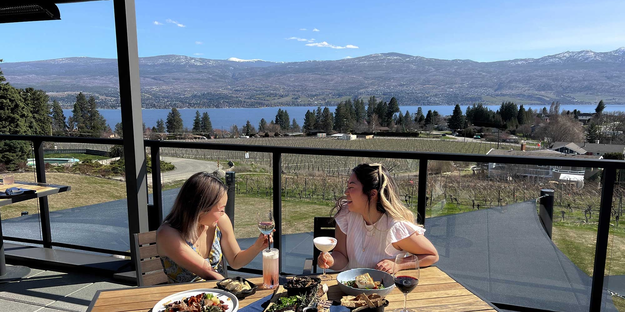 Emma Choo and Priscilla Banh sit at a table on a patio on a sunny day. There are several dishes of food on the wooden table and views of the lake behind them.