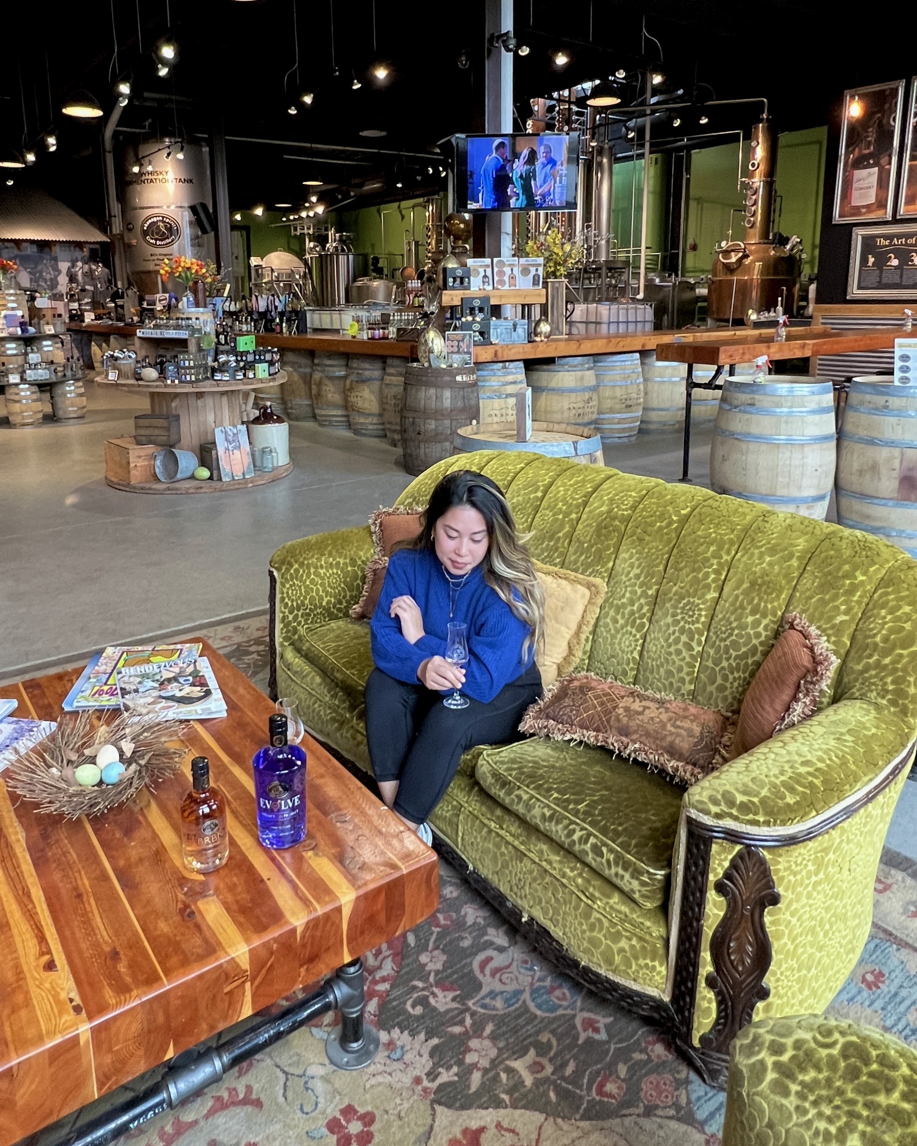 A woman wearing a blue jacket sits on a large green couch at Okanagan Spirits Craft Distillery. She is holding a glass of wine. In front of her is a low wooden table with other bottles on it.