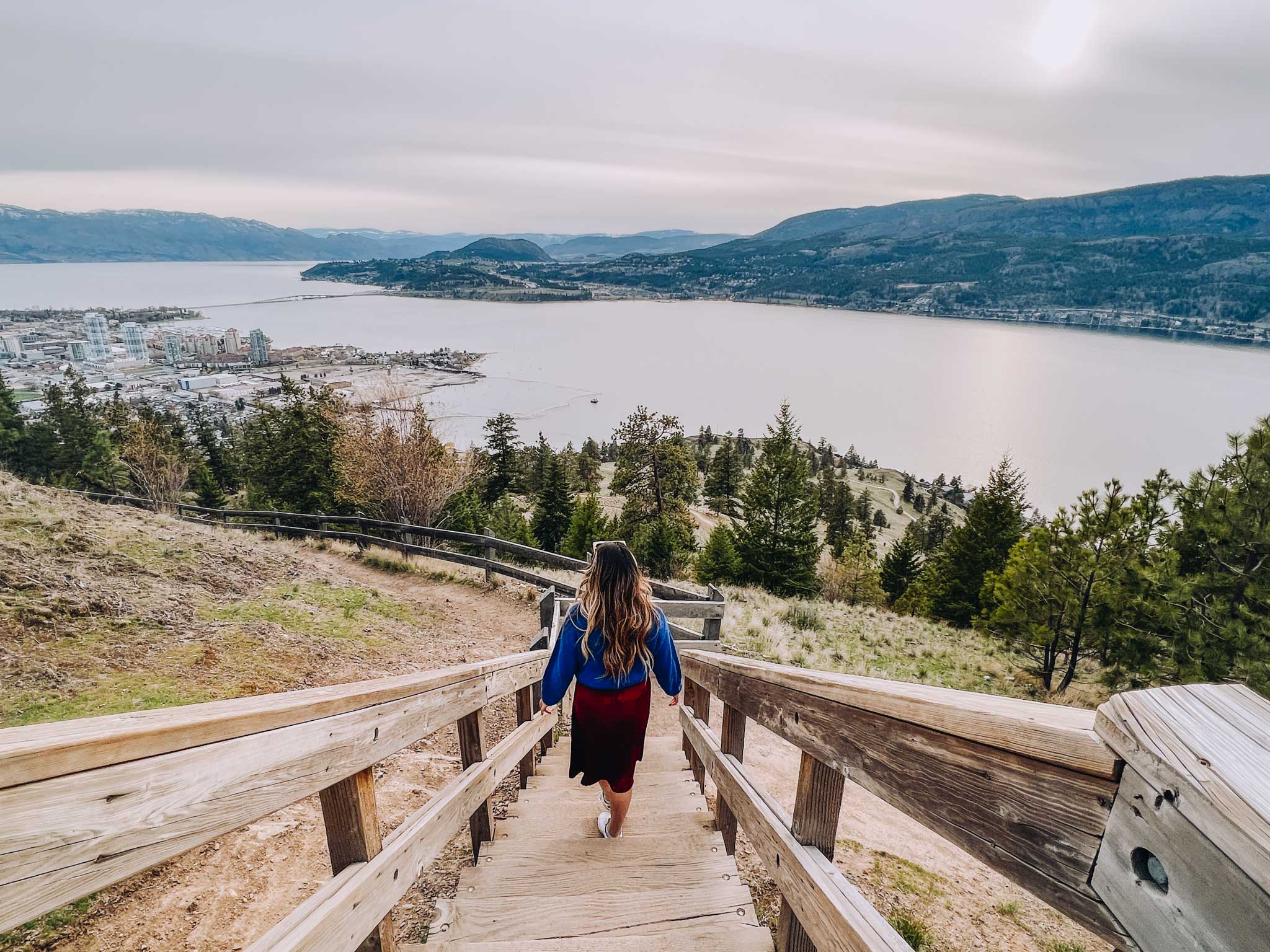 Emma Choo walks down a series of wooden stairs as she walks down from Knox Mountain. The stairs overlook the lake and mountain below. Emma is wearing a blue top and and a red skirt.