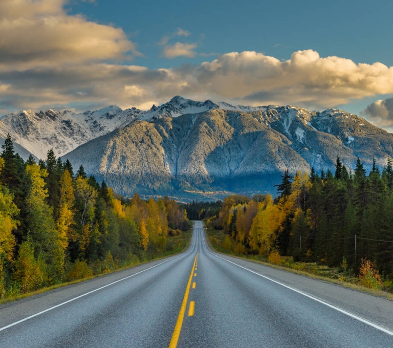 A highway runs toward snowy mountain peaks with dense forest on either side. There are a few clouds in a blue sky.
