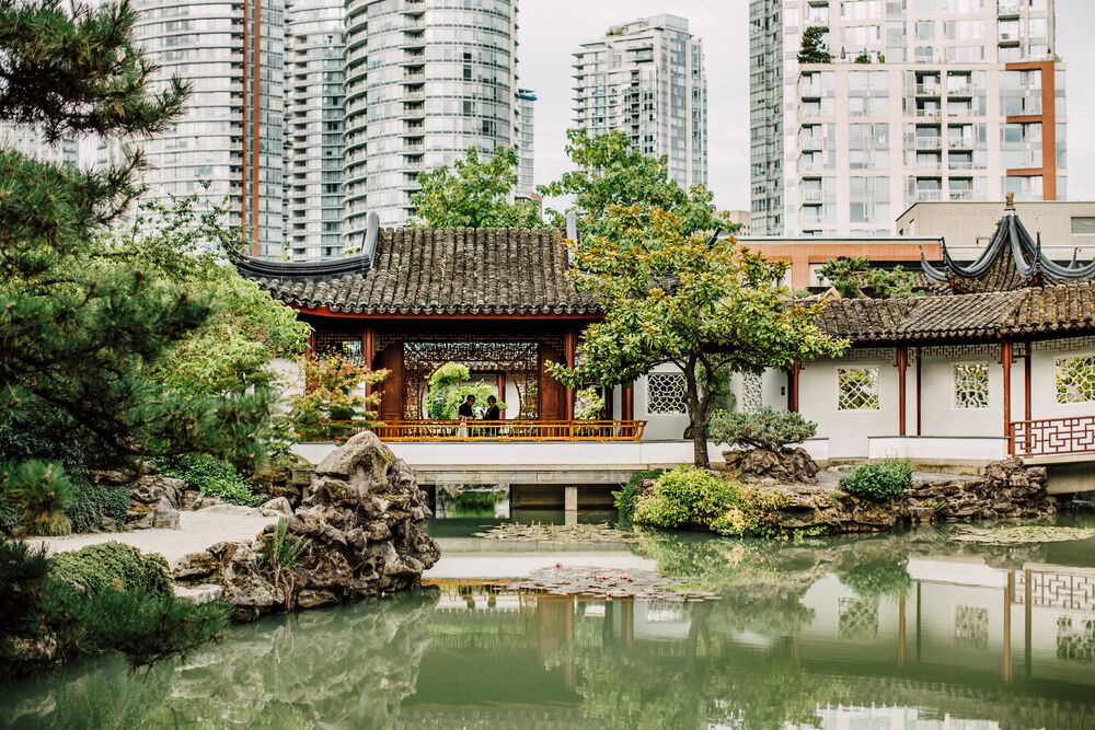 A green pond in the foreground is surrounded by a handful of evergreen trees with a traditional Chinese building behind, and in the distance the towers of downtown Vancouver.