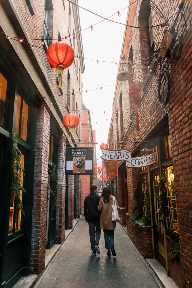 a couple walks down a very narrow alley with brick buildings on either side , and lanterns and strings of lights above.