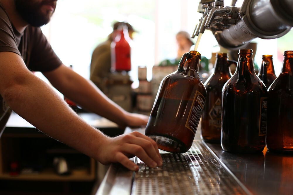 Two arms hold a tilted growler being filled with beer on tap. Empty growlers sit behind.