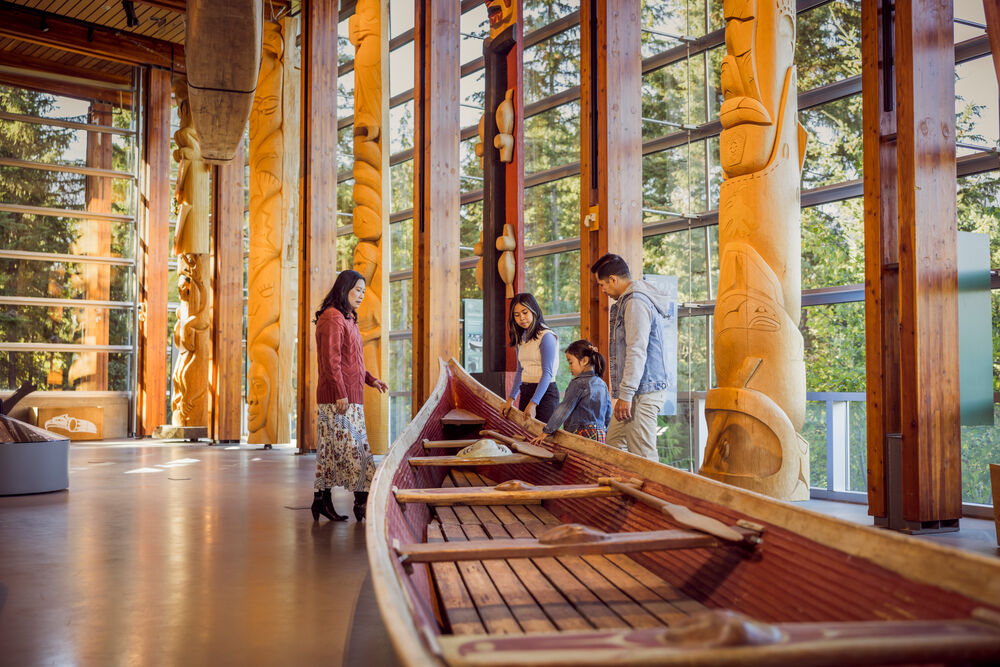 A family of four looks at an Indigenous canoe and carved totem poles at Squamish Lil'wat Cultural Centre.