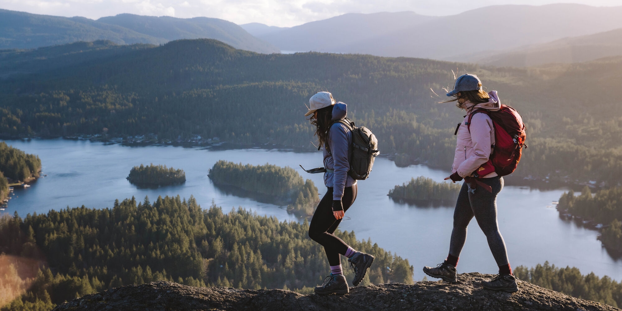 Two women wearing baseball caps and backpacks, walk along a rocky mountaintop overlooking forested hills with a narrow body of water in the middle of the frame containing a few forested islets