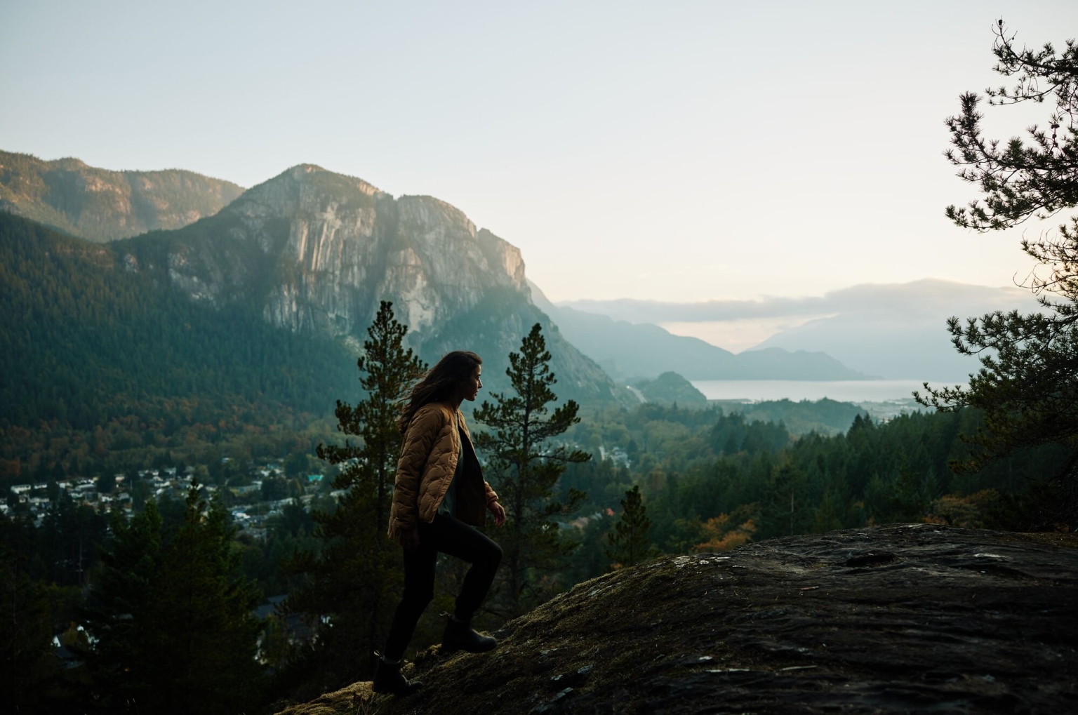 A person hikes along a granite rockface with views of Howe Sound and the Stawamus Chief monolith.