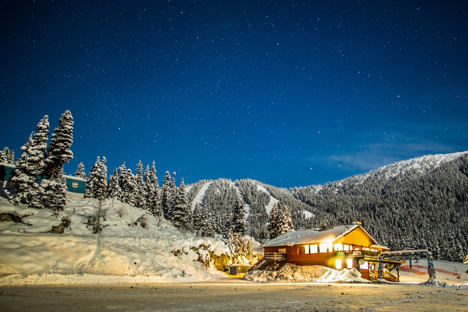 A wooden lodge sits in a gorgeous snowy setting with trees and mountains behind. It's dusk, and the lights are on inside the lodge.