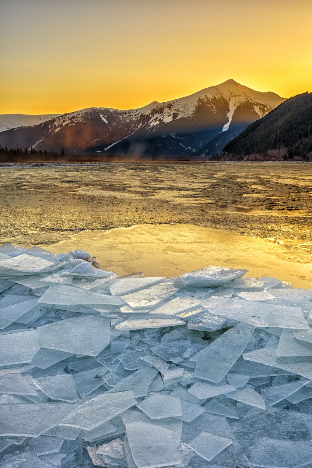 Thin squares of ice sit piled up on the surface of a lake at sunrise, mountains in the background.