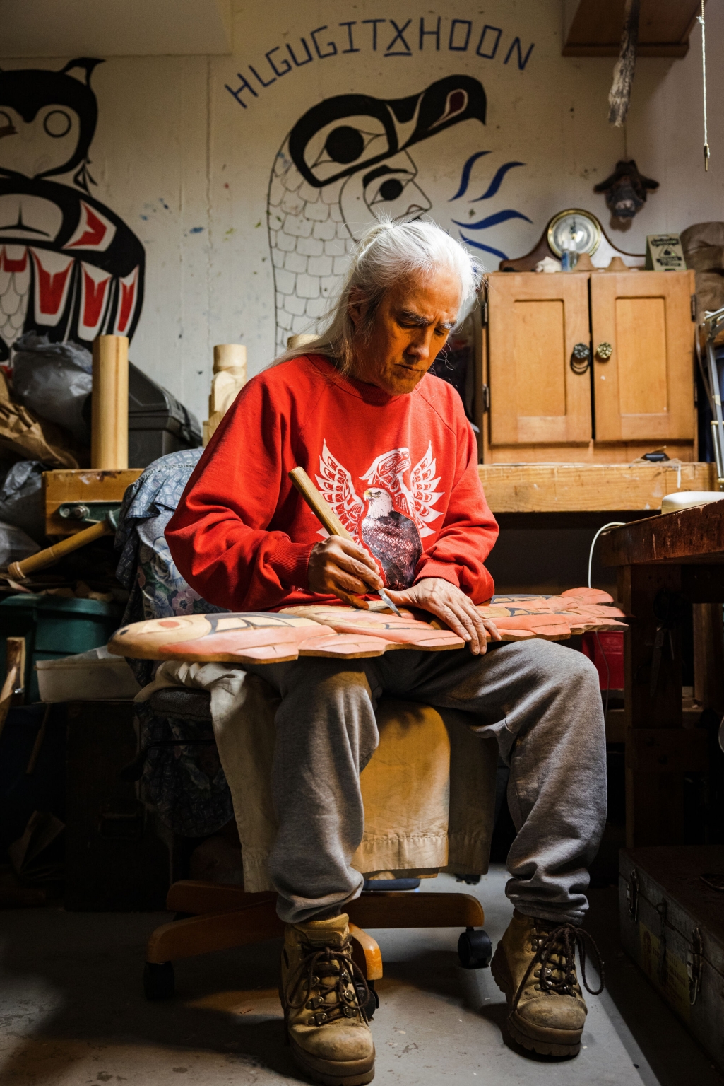An Indigenous artist in a red sweatshirt sits in a chair in what looks to be his workshop working on a long, flat carving.