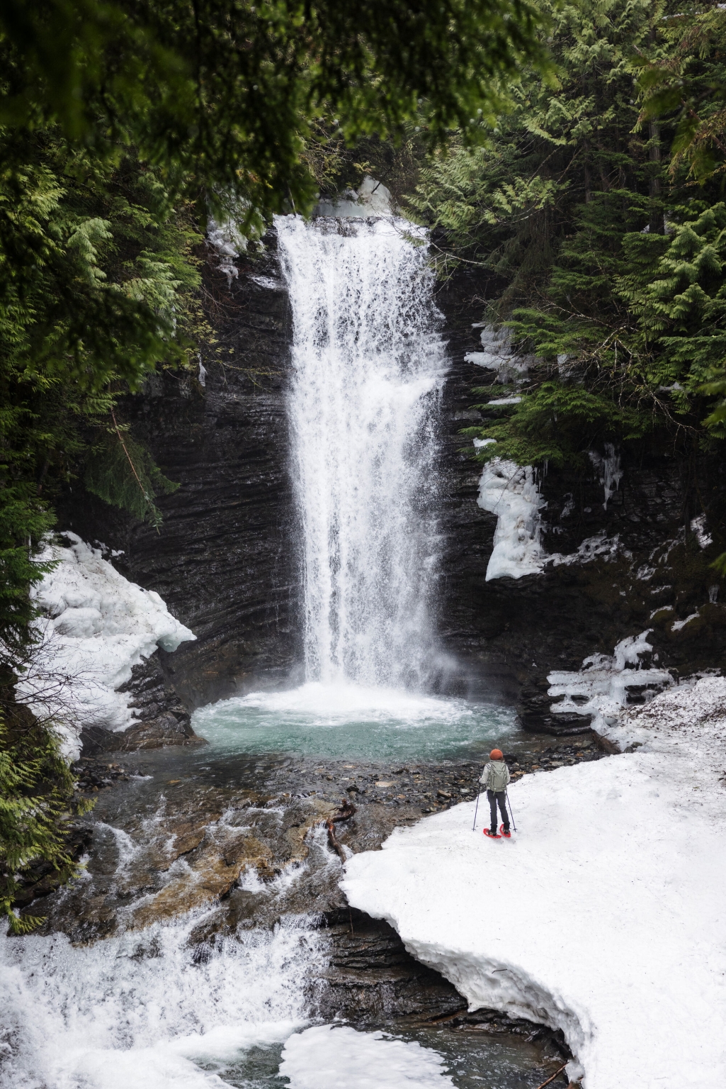 A person stands on the snowy banks of a body of water as a short waterfall plummets into a pool.