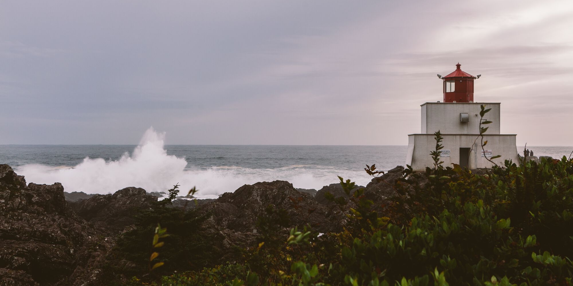 A short white lighthouse with a red top sits on the right side of the image o a rocky shorline with waves breaking in the background.