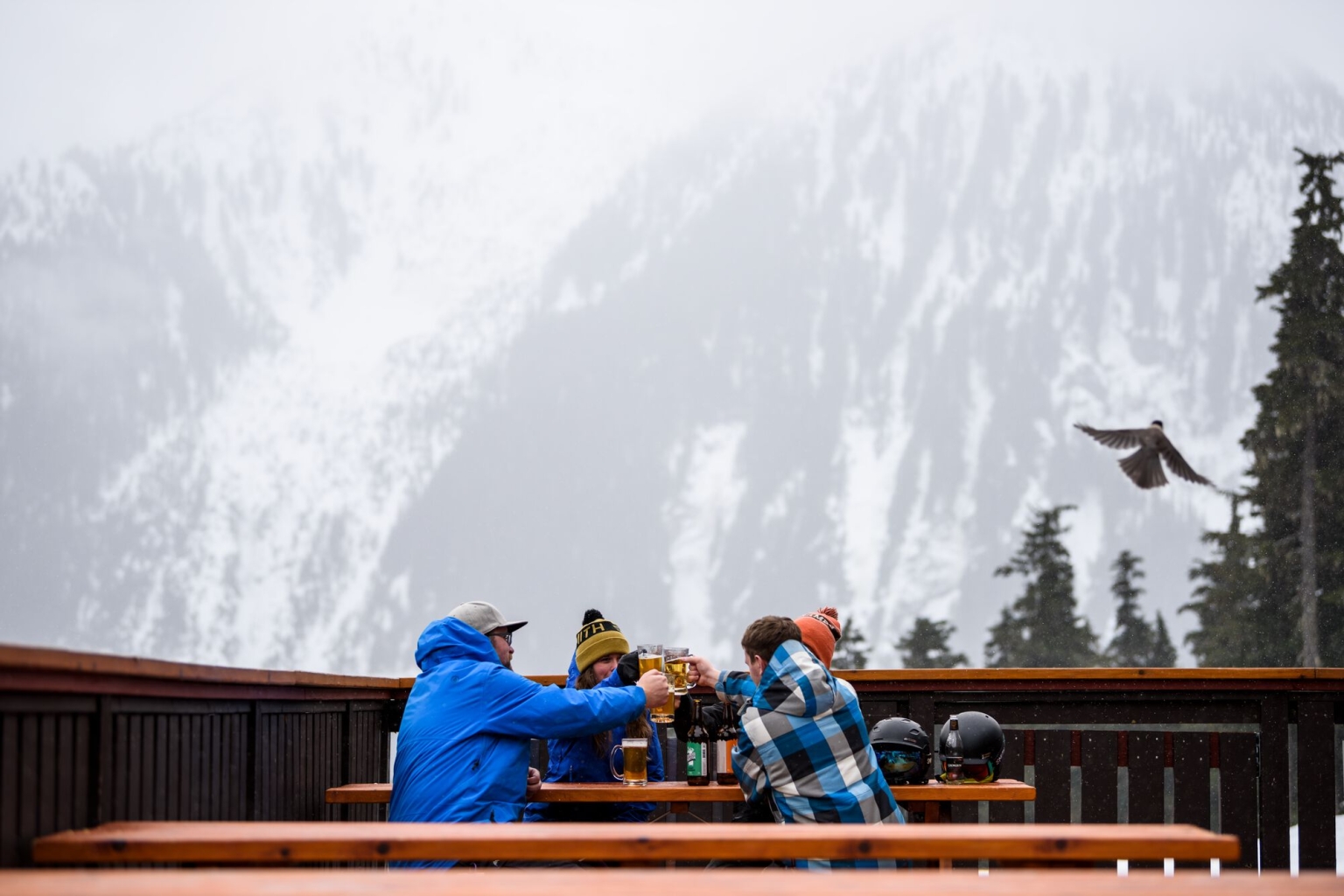 Four people in ski attire sit at a wooden picnic table on an outdoor patio with a snow-covered mountain behind them slightly obscured by fog. The four are clinking beer glasses.