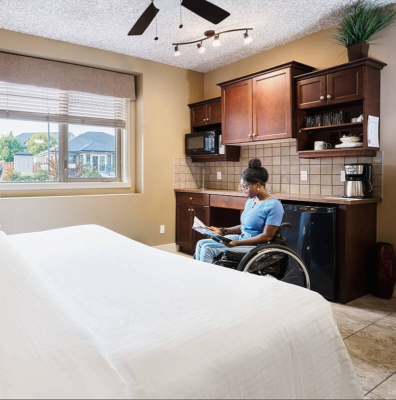 A person in a wheelchair reads a pamphlet at the foot of a bed in a hotel room.