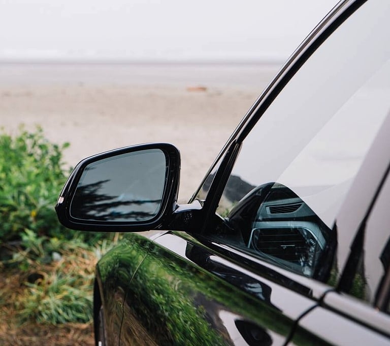 A black car is parked in front of a sandy beach with green bushes on the left of the image.
