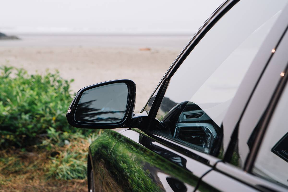 A black car is parked in front of a sandy beach with green bushes on the left of the image.